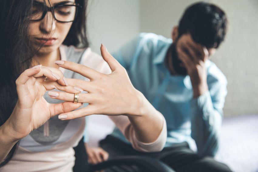 Junge Frau nimmt Ehering vom Finger, Mann sitzt unglücklich im Hintergrund - Was tun mit Ehering nach der Scheidung?